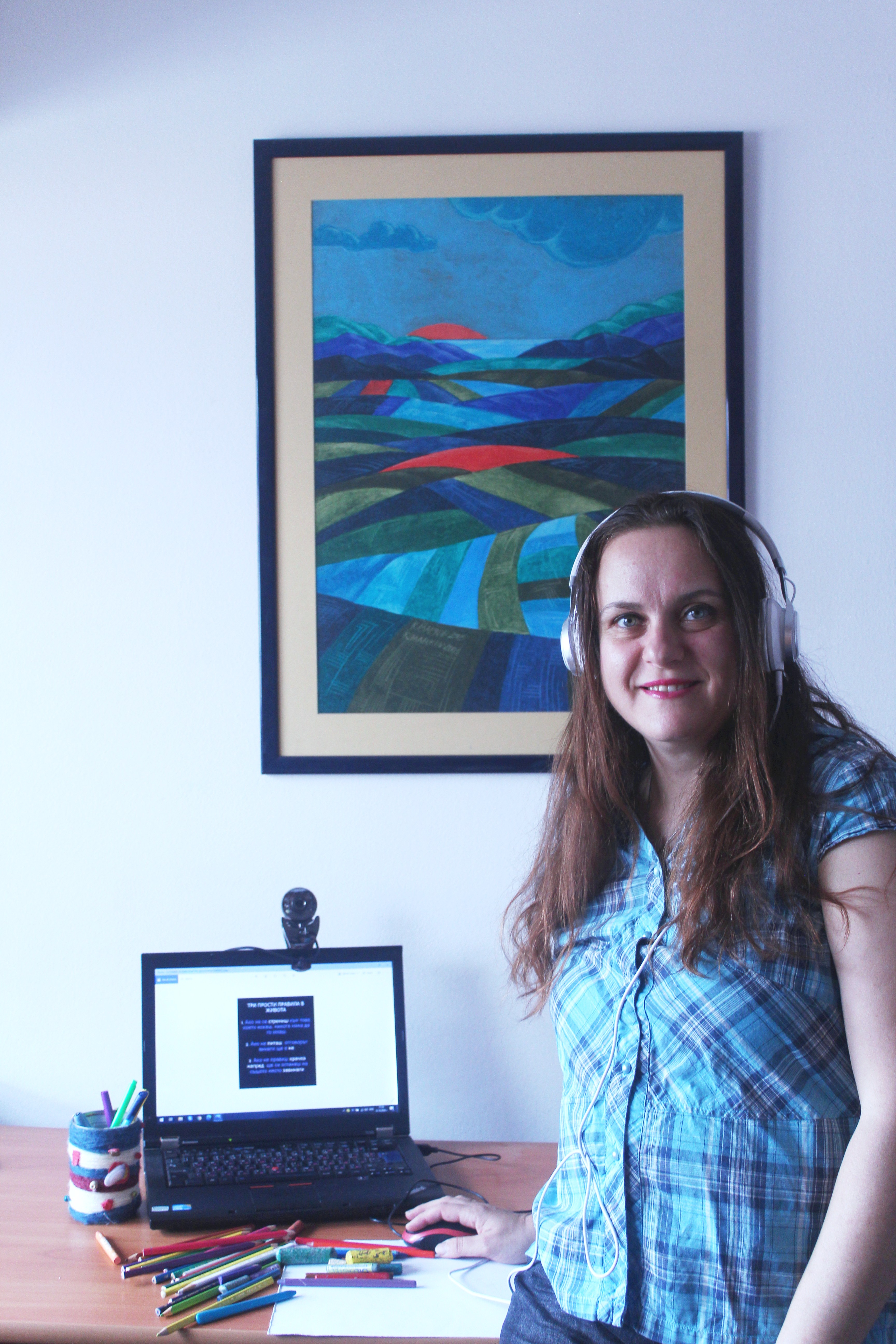 Това е изображение на млада жена пред компютър и арт материали, на фона на красива картина