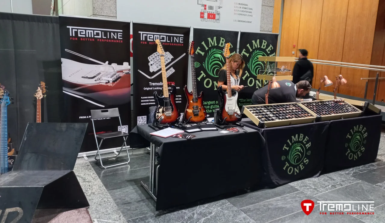 <img src=”Tremoline-GuitarSummit2025-1280x741-v02a” width="1280" height="741" alt=” Two people near to guitar stand” />